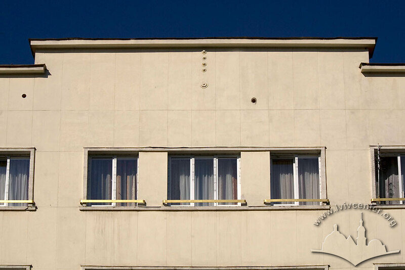 Просп. Свободи, 5. Центральна частина фасаду в рівні IV-го пов. з мінімалістичним віконним обрамленням і жардиньєрками та аттиком